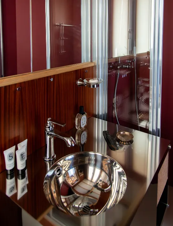 Vasque ronde en inox miroir entourée de produits d'accueil et d'un peigne, dans une salle d'eau aux murs bordeaux et lambris de bois.
