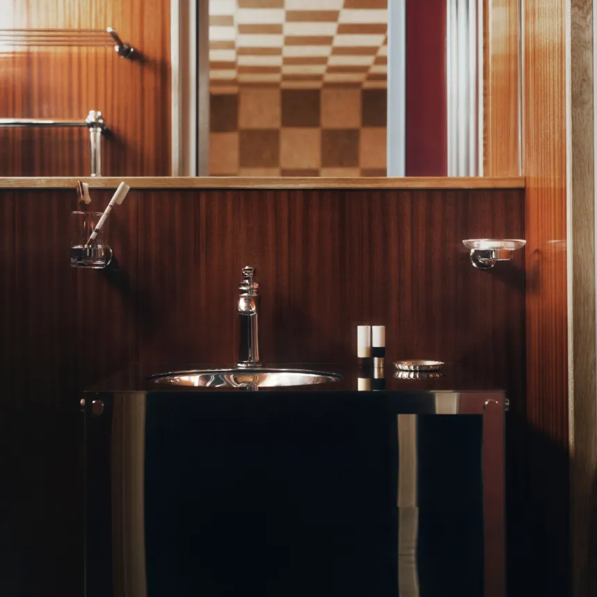 Lavabo en métal poli sur un meuble noir laqué, devant un lambris d'acajou et un miroir reflétant un plafond à damier.