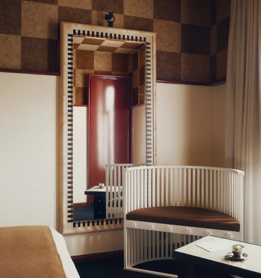 Fauteuil à claire-voie blanc et assise marron devant un miroir en pied au liseré graphique, reflétant une porte laquée rouge.
