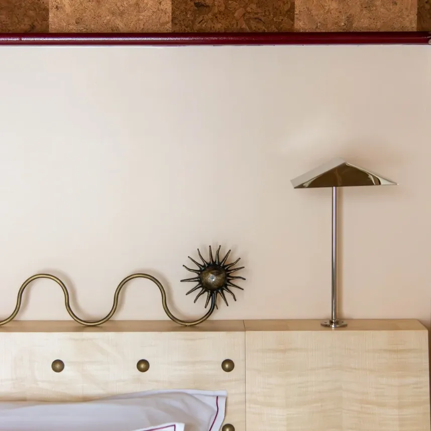 Détail d'une tête de lit en bois clair surmontée d'une sculpture ondulée en laiton et d'une lampe chromée, contre un mur rose poudré.