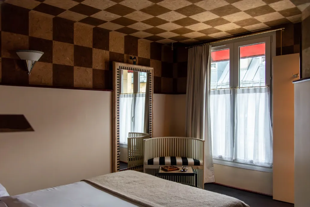 Vignette d'une chambre éclairée par une fenêtre aux rideaux blancs, avec un fauteuil clair et un décor en damier brun au plafond.
