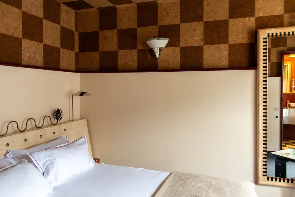 Petite vignette d'une chambre aux murs crème, avec un lit blanc à gauche, une applique conique et une frise de liège à damier en hauteur.