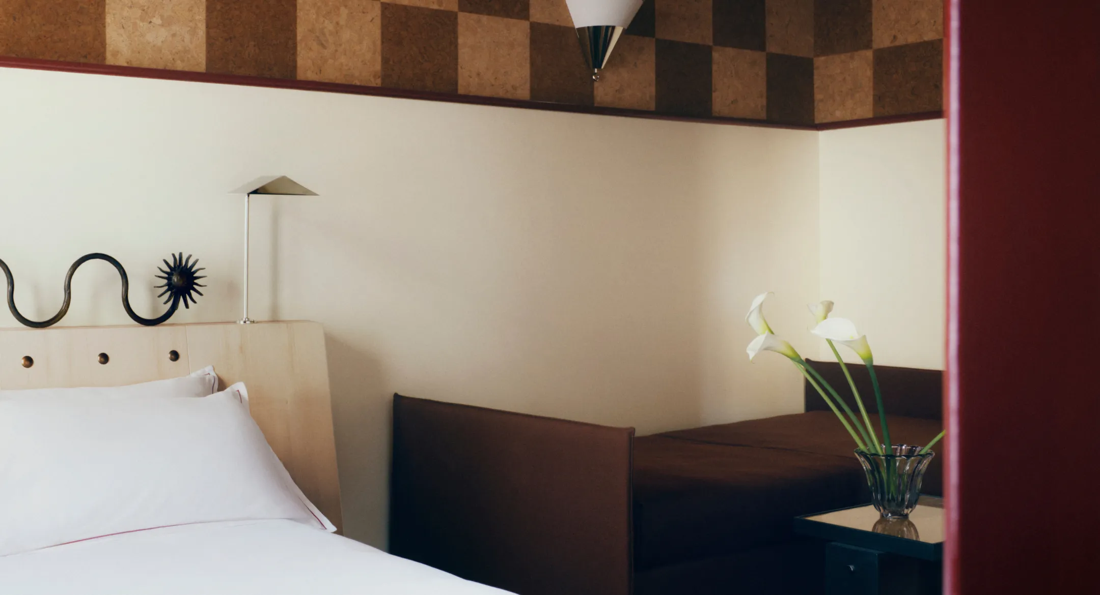 Coin salon d'une chambre avec une banquette en cuir brun, des arums blancs dans un vase en cristal et une tête de lit décorée d'une arabesque en fer noir.
