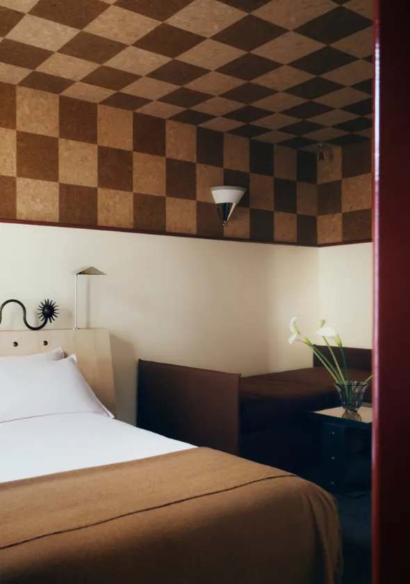 Chambre cadrée par l'encadrement d'une porte rouge, dévoilant un lit au plaid cognac, une banquette sombre et des murs coiffés d'un damier en liège.
