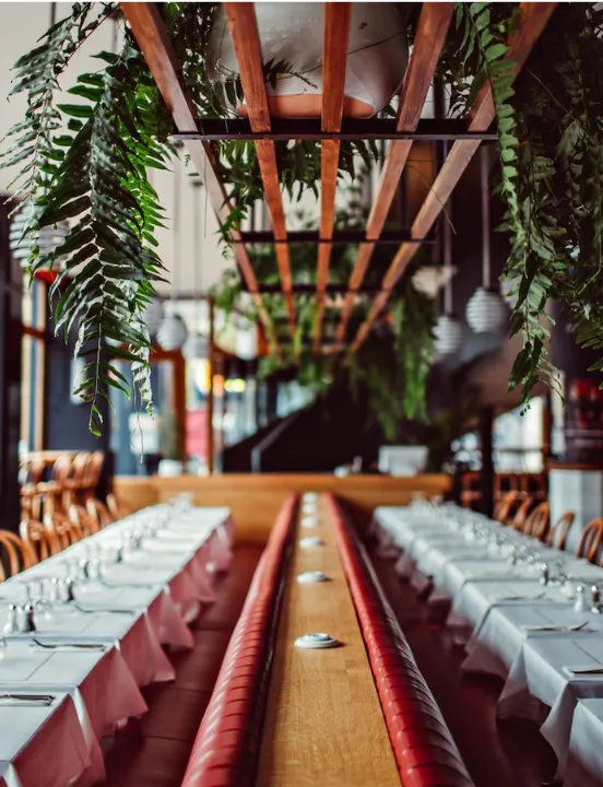Salle de restaurant avec deux rangées de tables nappées de blanc, une longue banquette rouge centrale et des fougères suspendues à une structure en bois.