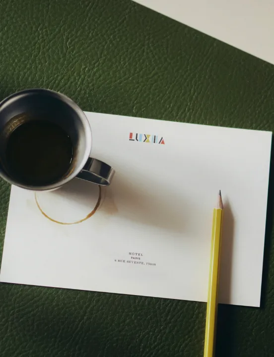 Carte à en-tête de l'hôtel Luxia posée sur un sous-main en cuir vert, avec une tasse de café en métal et un crayon jaune.