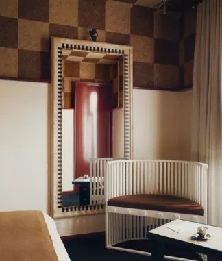 Coin d'une chambre avec un grand miroir encadré en damier fixé au mur, reflétant une porte rouge, un fauteuil blanc incurvé avec coussin brun, un petit table et un canapé beige partiel.