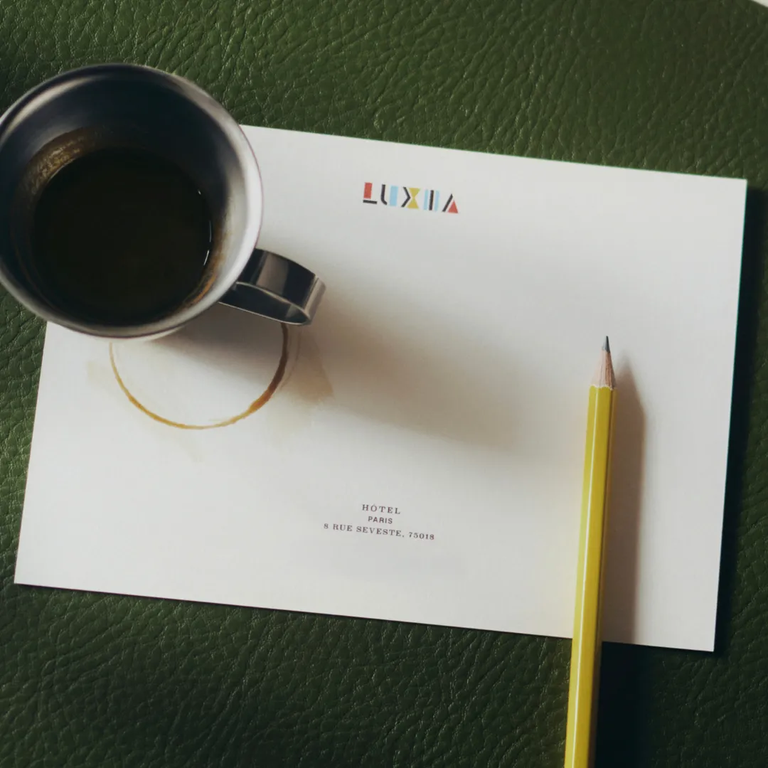 Une tasse en métal avec du café, posée sur une feuille blanche marquée Luxia et l'adresse Hôtel Paris 8 Rue Sevestre, avec une marque de tasse et un crayon jaune placé à côté.
