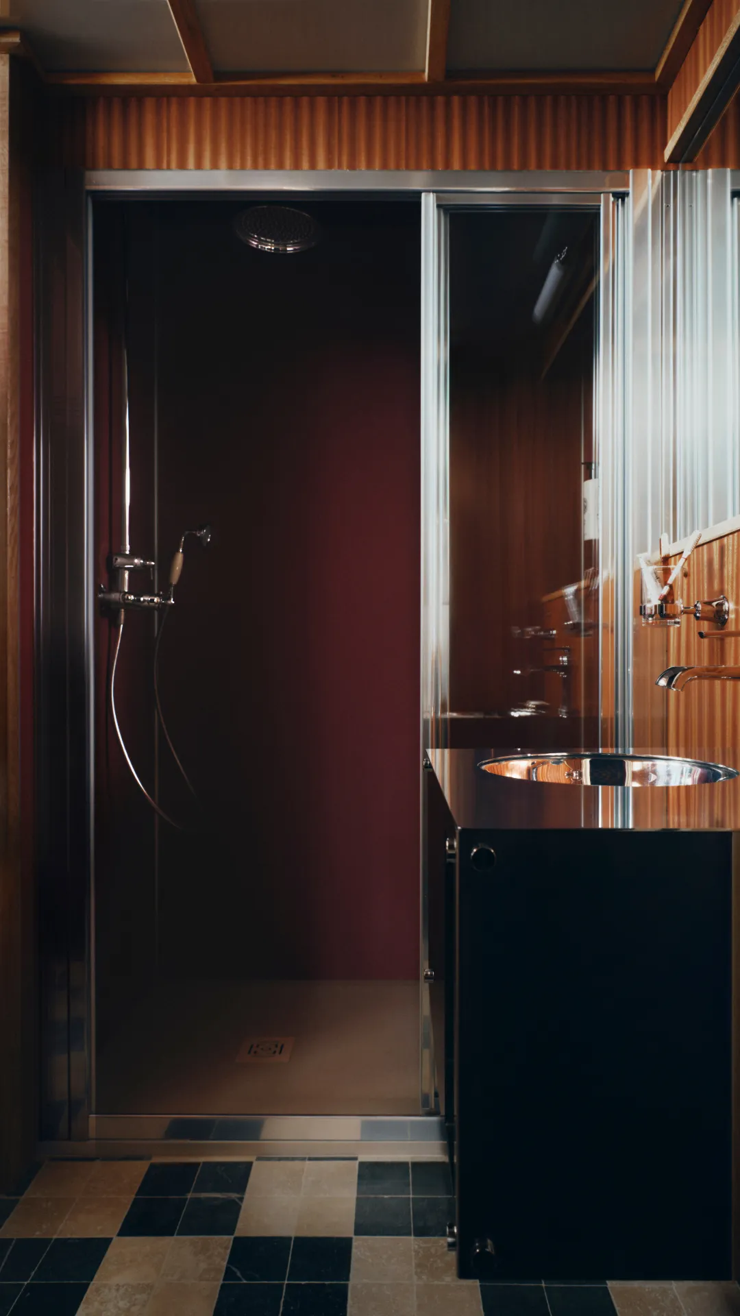Salle de bain moderne avec une cabine de douche à paroi vitrée, une pomme de douche et un lavabo rond encastré dans un meuble noir brillant avec un mur en bois à l'arrière-plan.