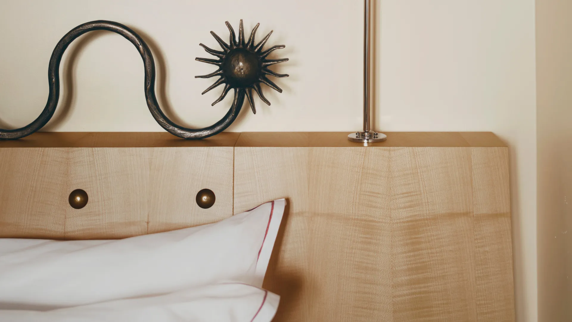 Chevet en bois clair avec une décoration en métal noir en forme de soleil à gauche et un luminaire métallique à droite, avec un oreiller blanc à bord rouge partiellement visible en bas.