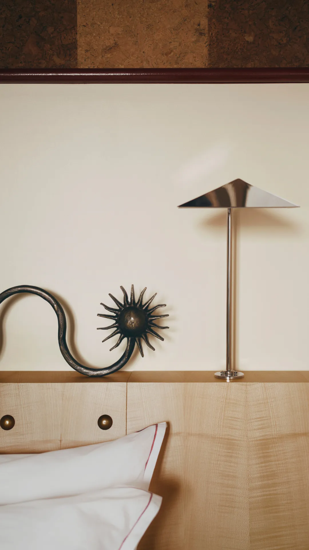 Tête de lit en bois clair avec deux chevets en métal sombre en forme de soleil et une lampe moderne en métal chromé au-dessus, avec des oreillers blancs bordés de rouge en bas à gauche.