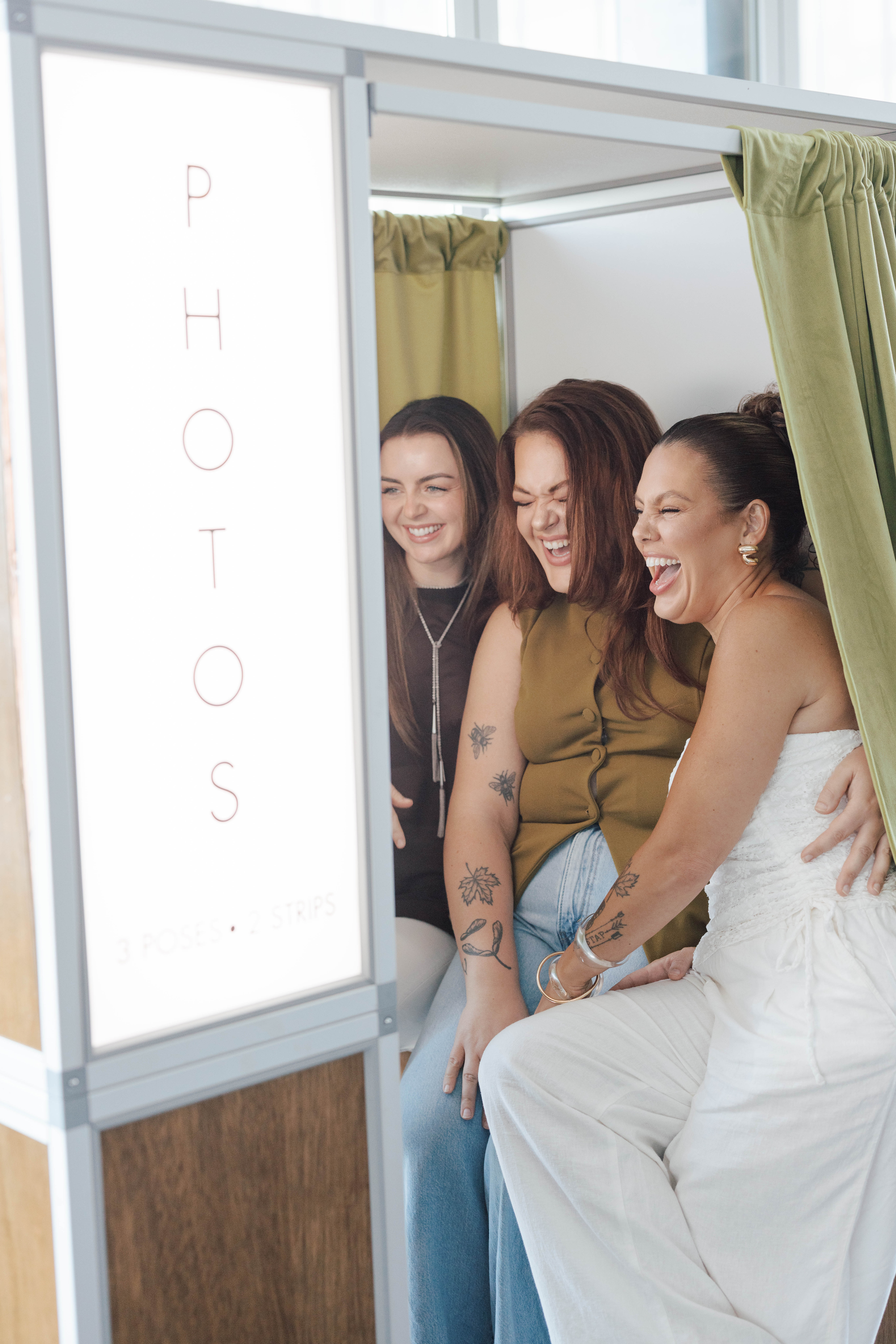 Three women sitting inside a photo booth laughing and smiling together with a green curtain.