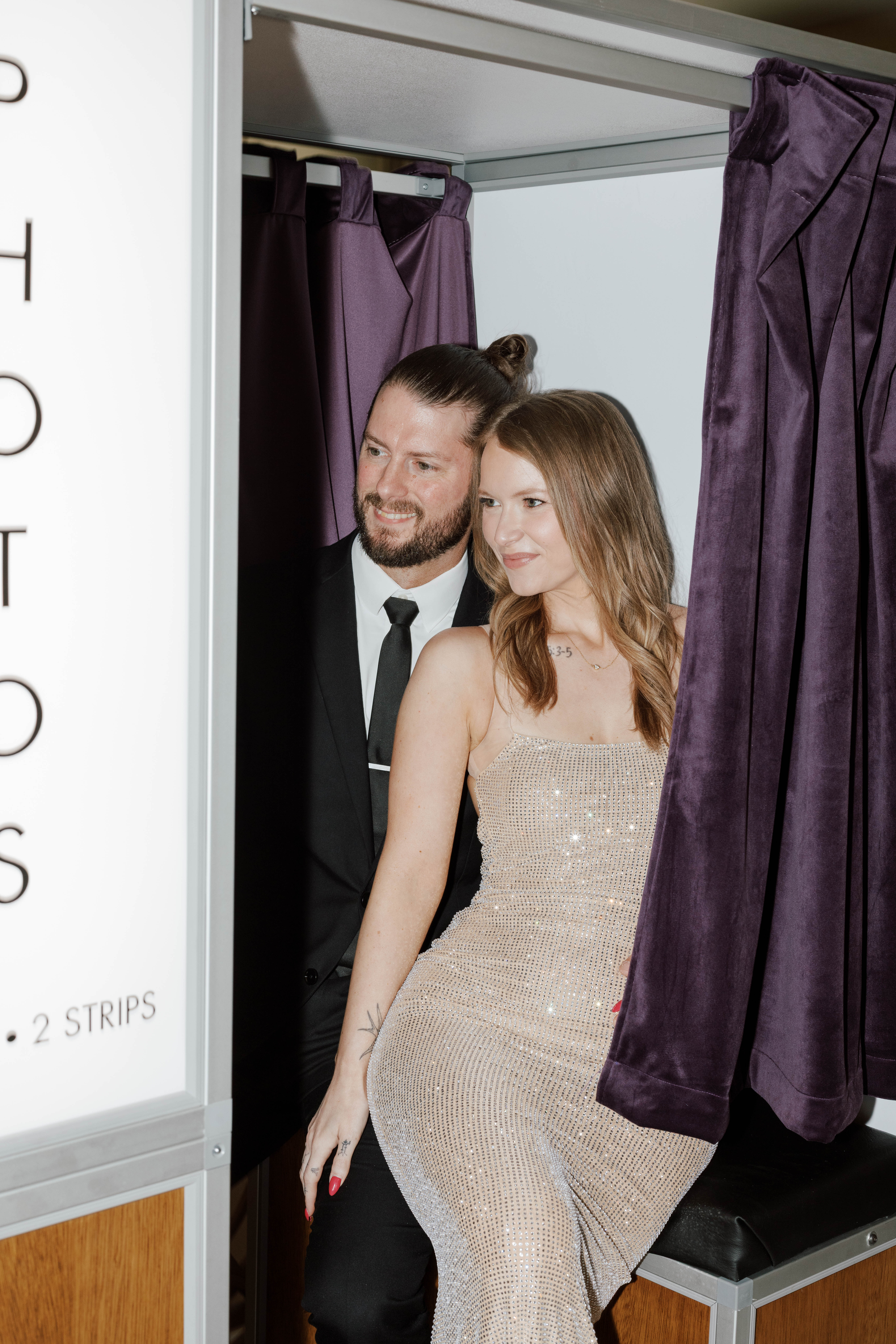 Couple dressed formally sitting inside a photo booth with purple curtains partially open.