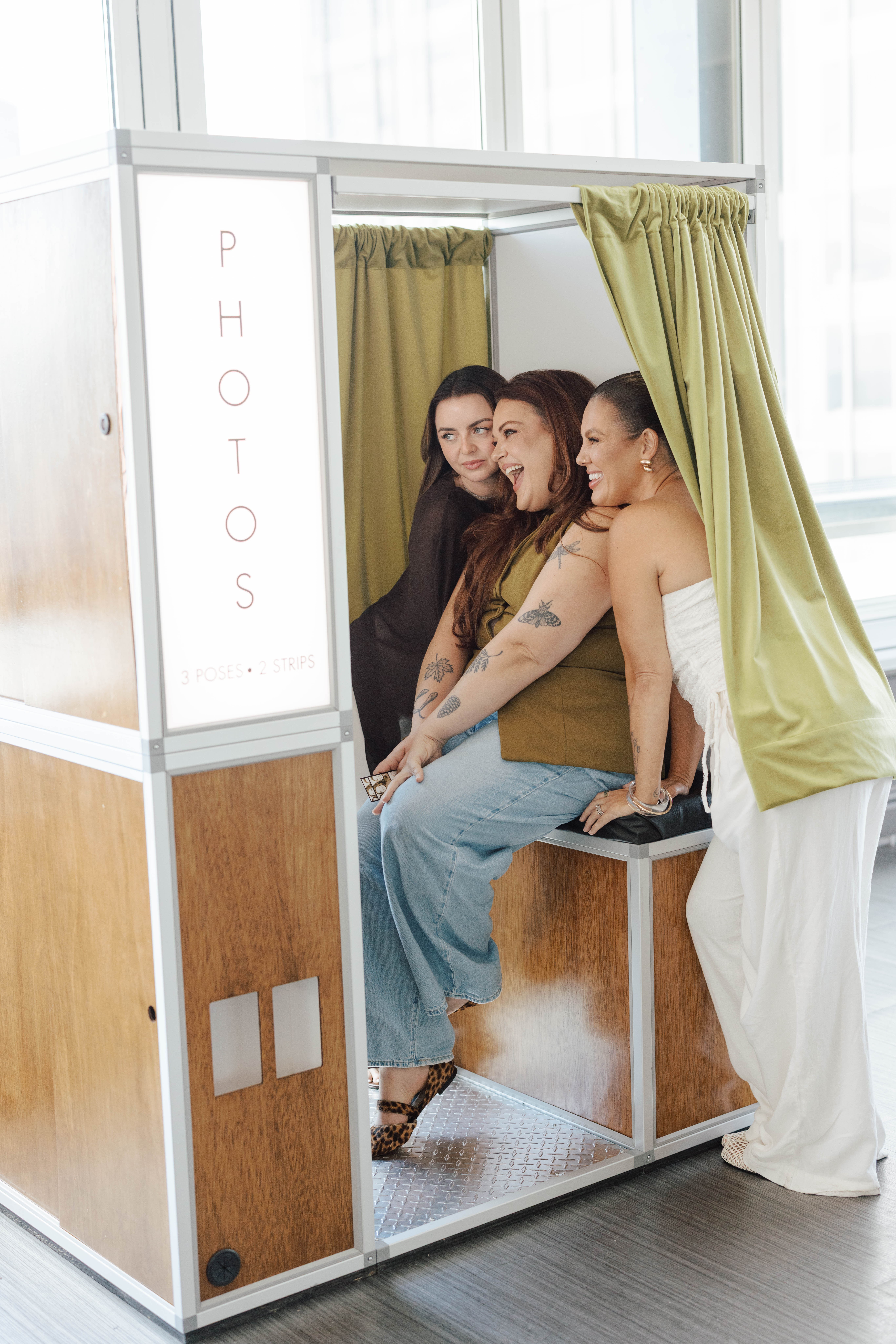 Three women smiling and posing inside a vintage-style photo booth with green curtains.