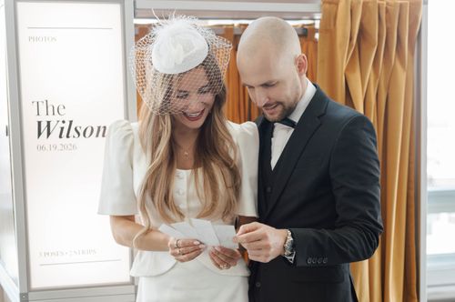 Smiling bride in white dress and groom in black suit looking at photo strip together indoors.