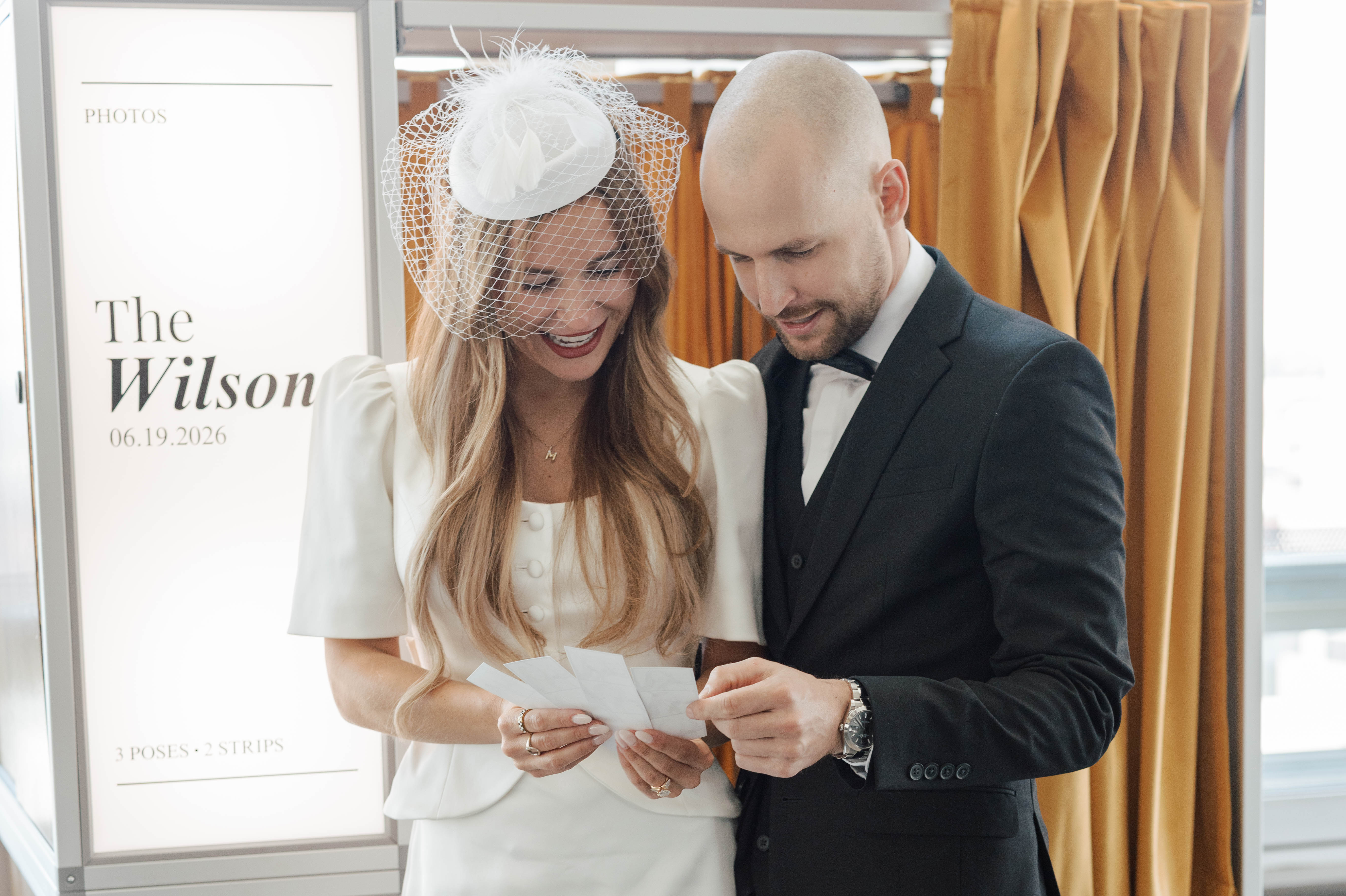 Smiling bride in white dress and groom in black suit looking at photo strip together indoors.
