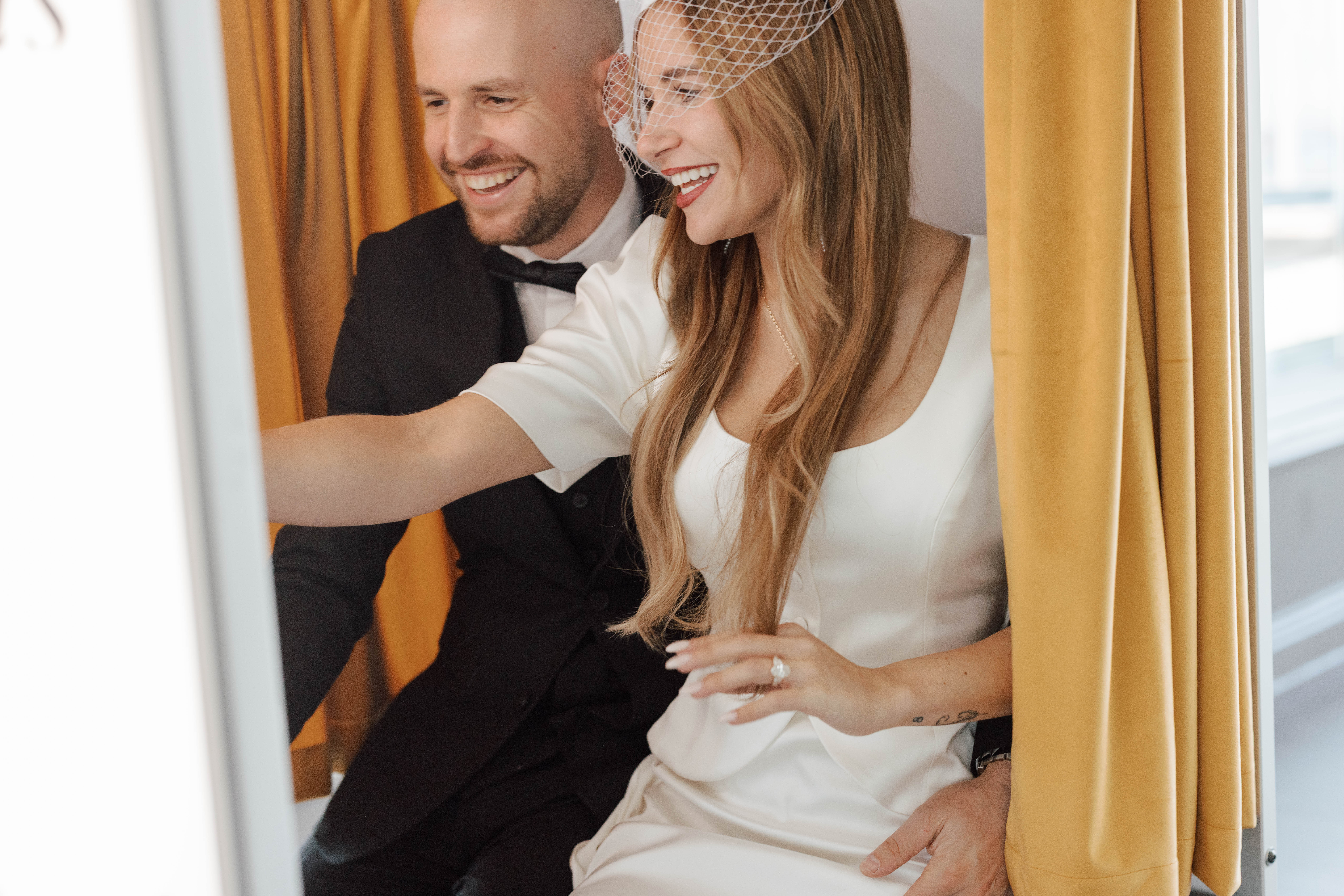 Smiling bride and groom dressed elegantly sitting close together behind mustard curtains.