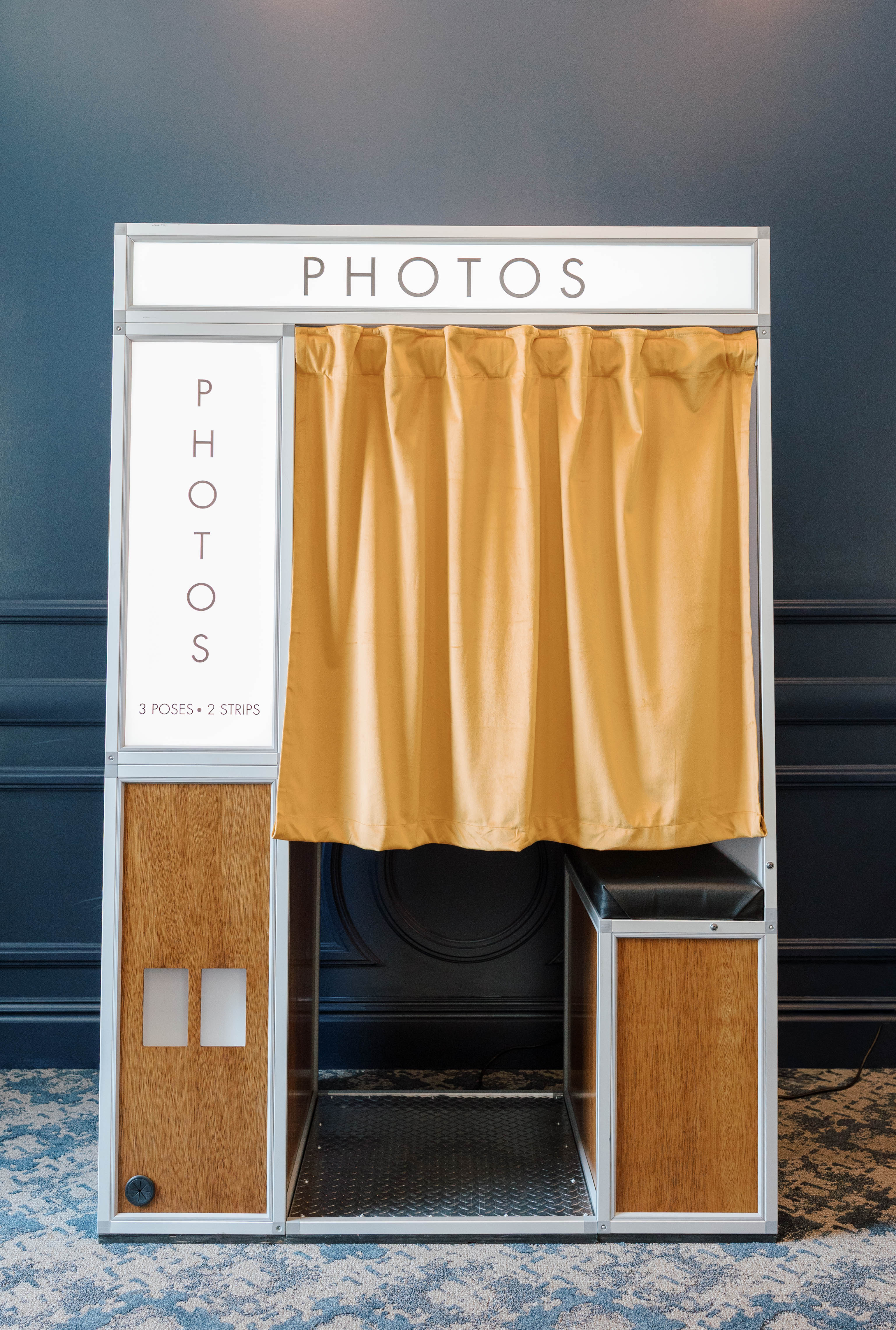 Vintage photo booth with mustard-colored curtain against a dark blue paneled wall and patterned carpet.