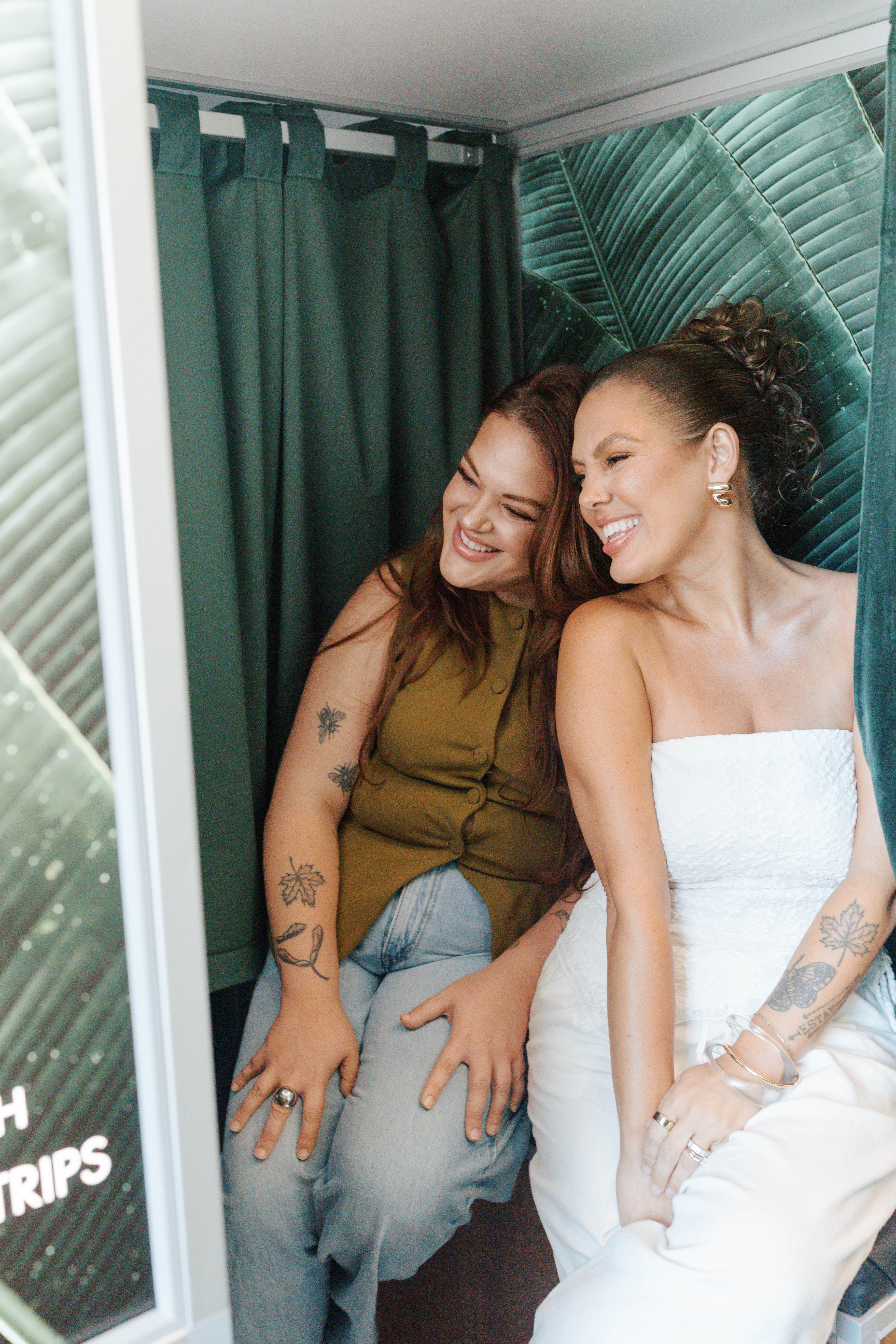 Two smiling women sitting closely in a small booth with green curtains and leaf-patterned walls.