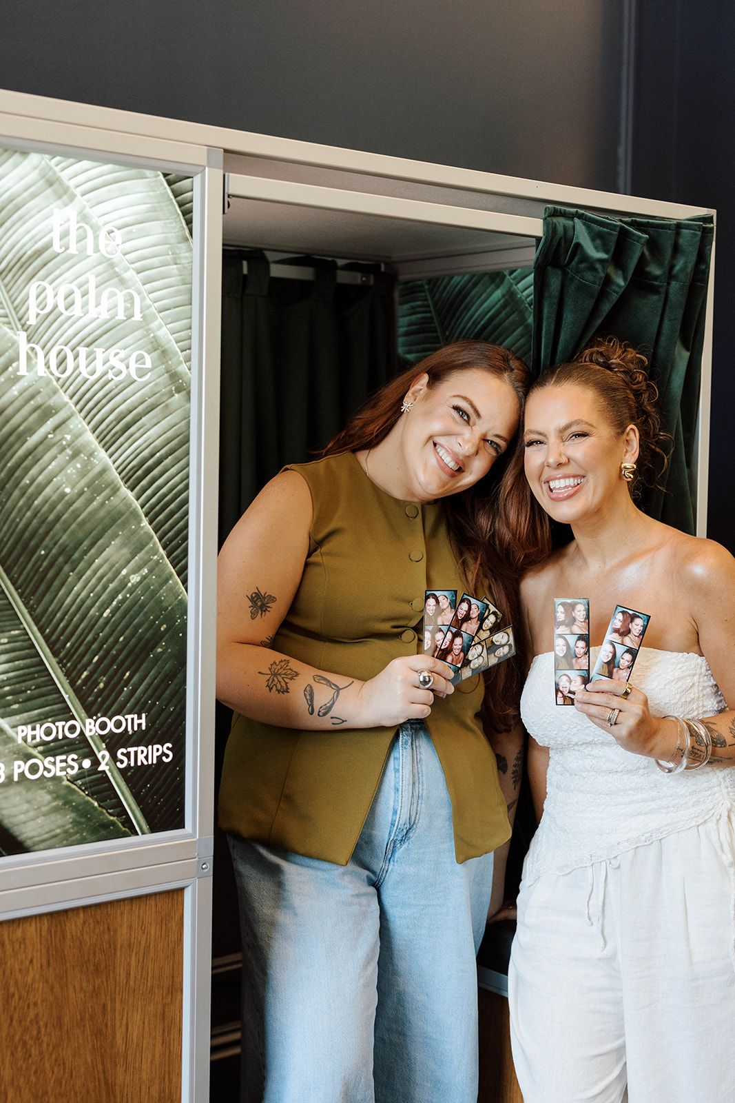 Two smiling women holding photo booth strips inside a photo booth with tropical leaf background.