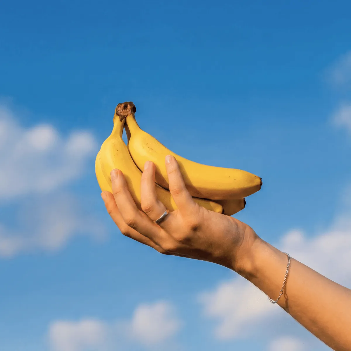 Hand wearing a ring and bracelet holding three ripe bananas against a blue sky with clouds.