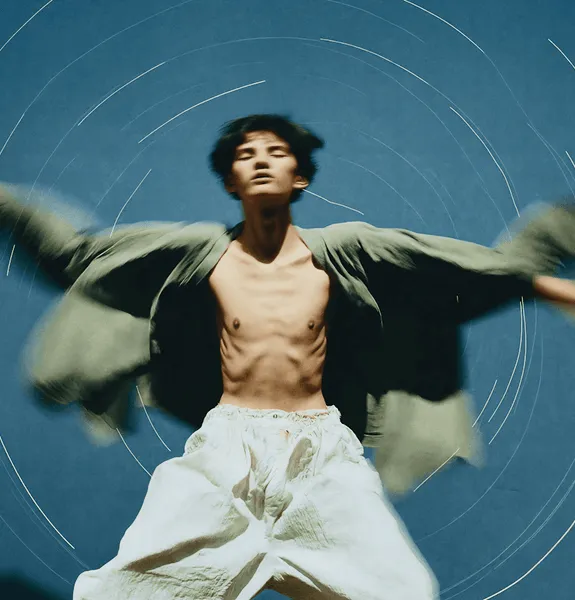 Artistic photo of a shirtless man in loose pants and open jacket with outstretched arms against a blue background.