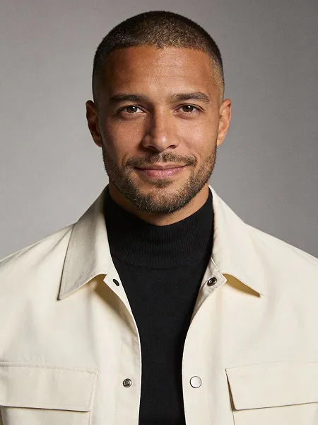 Smiling man with short hair and beard wearing a black turtleneck and light-colored jacket.
