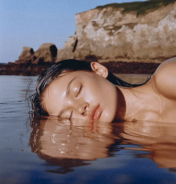 Woman with wet hair resting her face peacefully on calm water near rocky cliffs under clear sky.