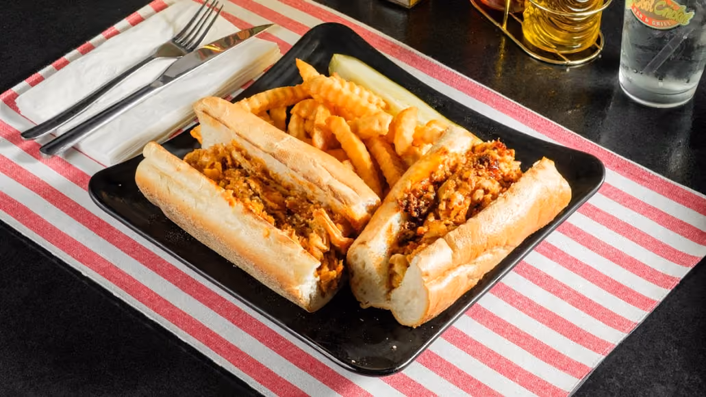 Plate with two chicken po' boy sandwiches, crinkle fries, and a pickle spear on striped cloth.