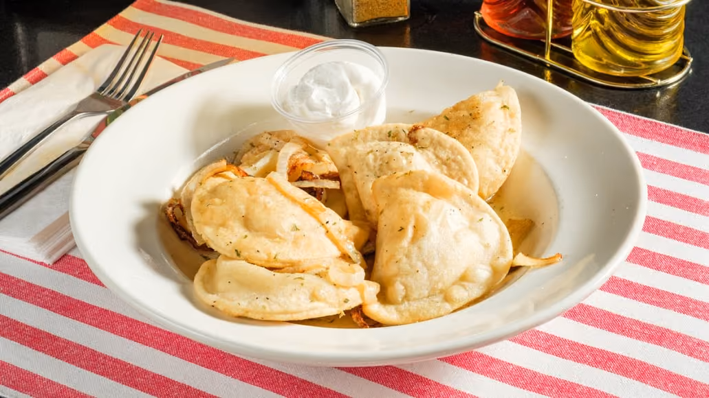 Plate of fried pierogi with grilled onions and a small cup of sour cream on a red-striped cloth.