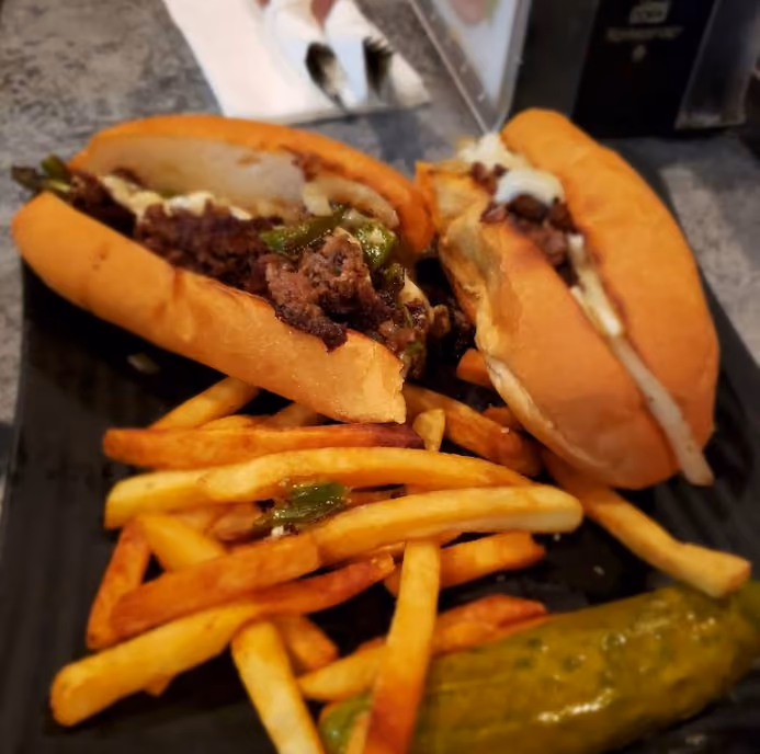 Two cheesesteak sandwiches with fries and a pickle on a black plate on a gray countertop.