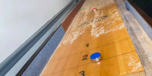 Shuffleboard table with blue and red pucks on a worn wooden surface marked with scoring zones.