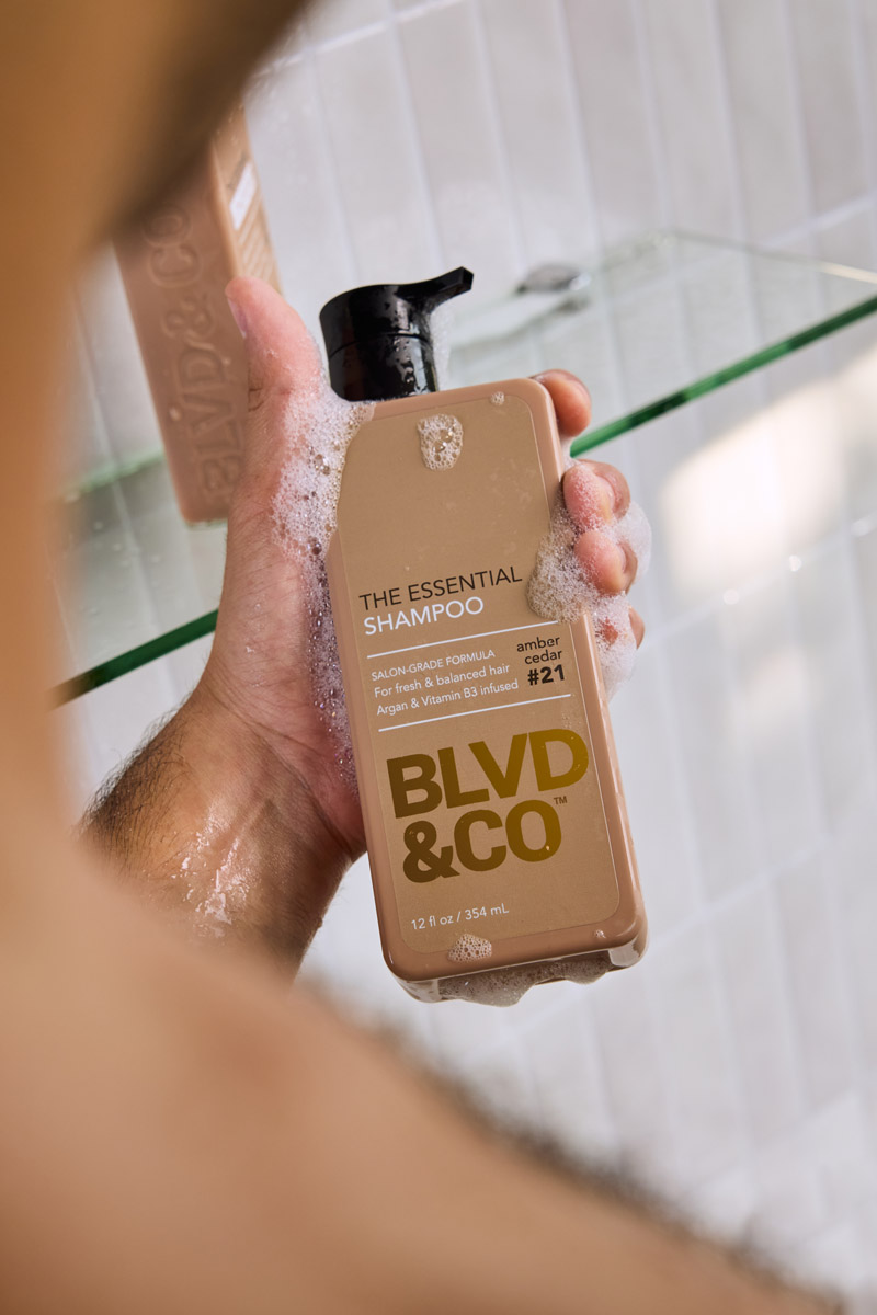 Hand holding a lathered bottle of BLVD&CO The Essential Shampoo in the shower