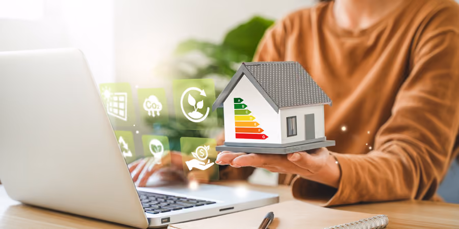 Person hält ein Modellhaus mit Energieeffizienzskala vor einem Laptop mit Symbolen für erneuerbare Energie, CO2-Reduktion und Einsparungen.