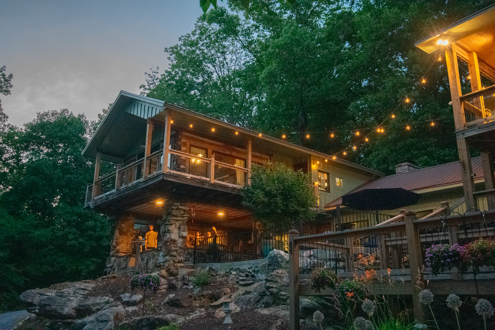 Rustic house with stone pillars, string lights, and trees at dusk with people on porch.
