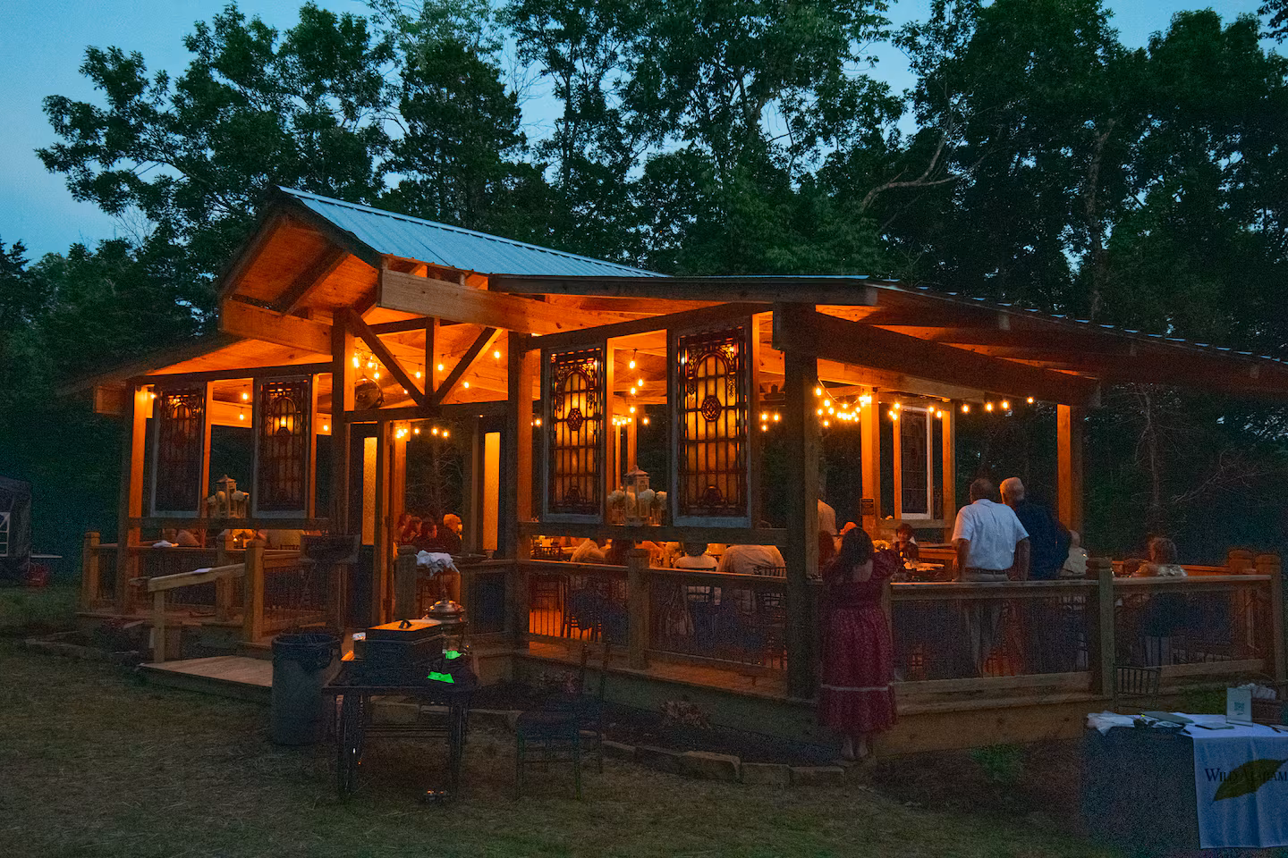 Wooden outdoor pavilion lit with string lights at dusk with people gathered inside and around it.