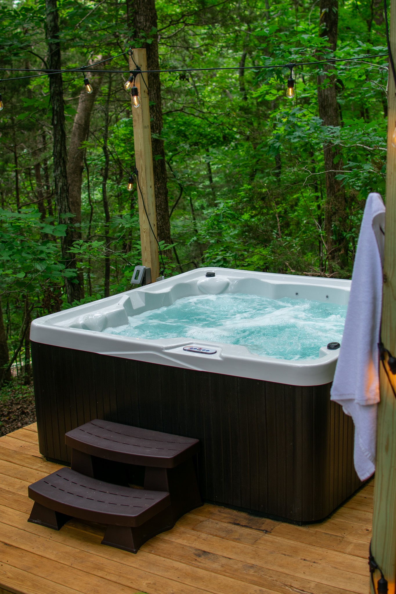 Outdoor hot tub with bubbling water, brown steps, and string lights in a green wooded area.
