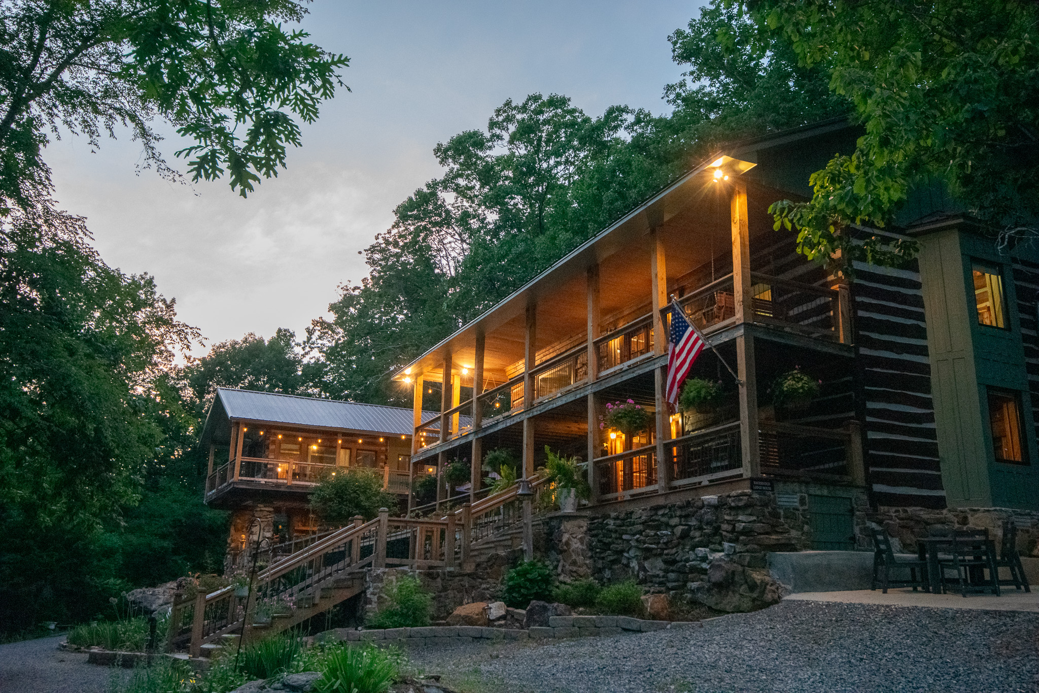Wooden lodge with lit porch lights and American flag, surrounded by trees at dusk.