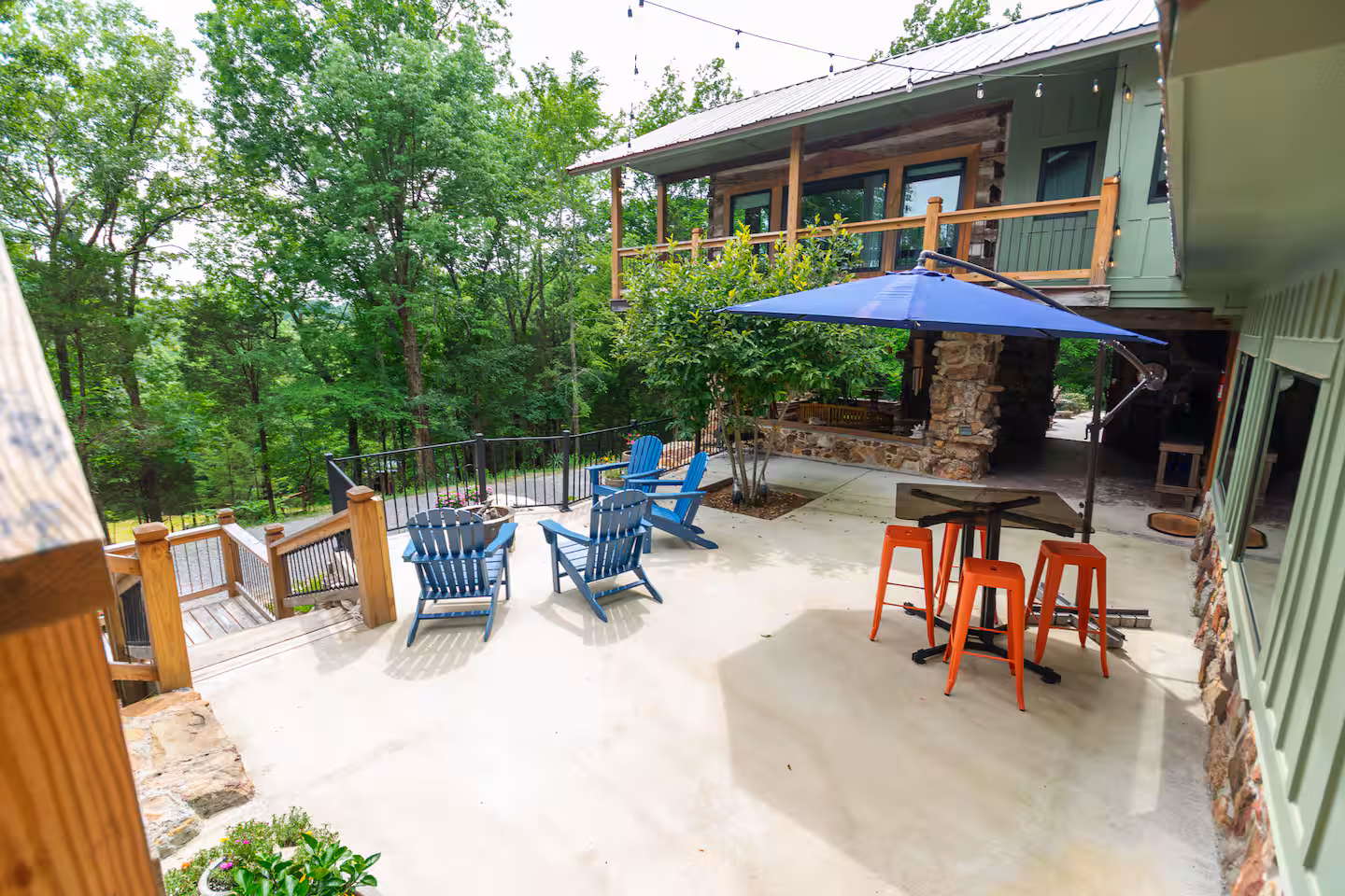 Patio with blue Adirondack chairs, orange stools under a blue umbrella, next to a green house and trees.