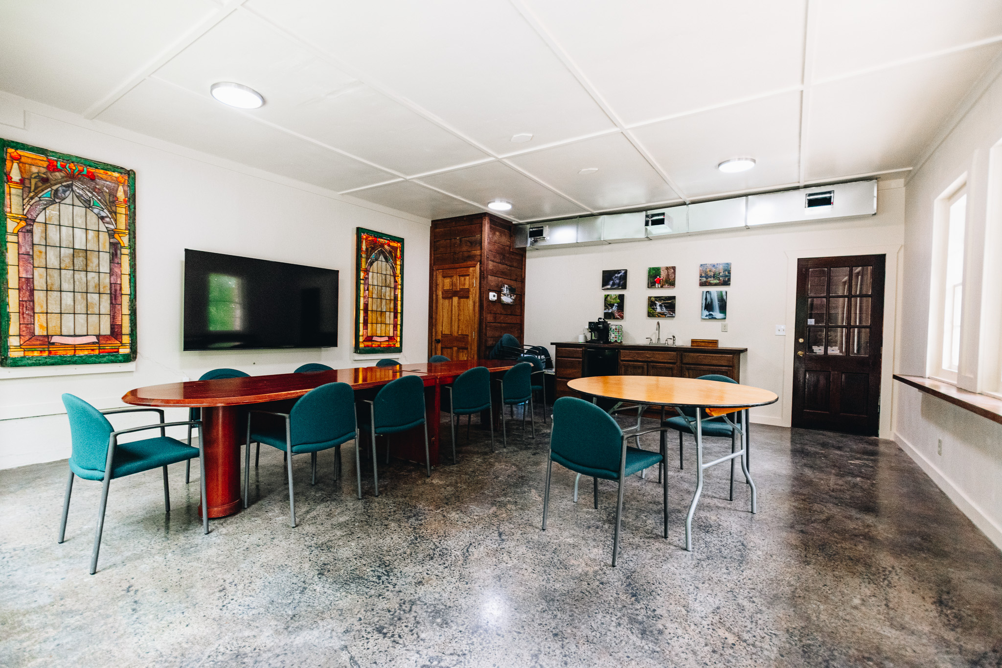 Conference room with two tables, teal chairs, stained glass windows, TV, and photos on far wall.