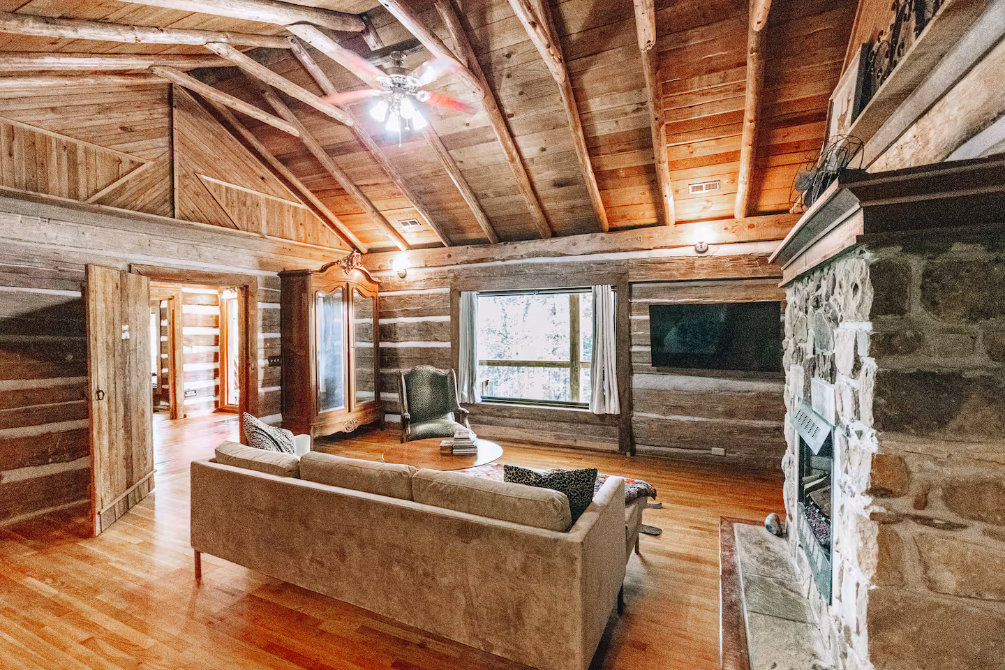 Cozy rustic living room with wooden beams, stone fireplace, beige couch, armchair, and a wall-mounted TV.
