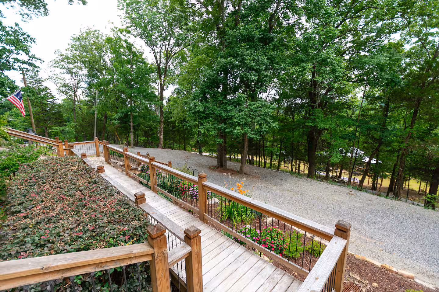 Wooden walkway with railing beside flowering plants and gravel driveway surrounded by green trees.