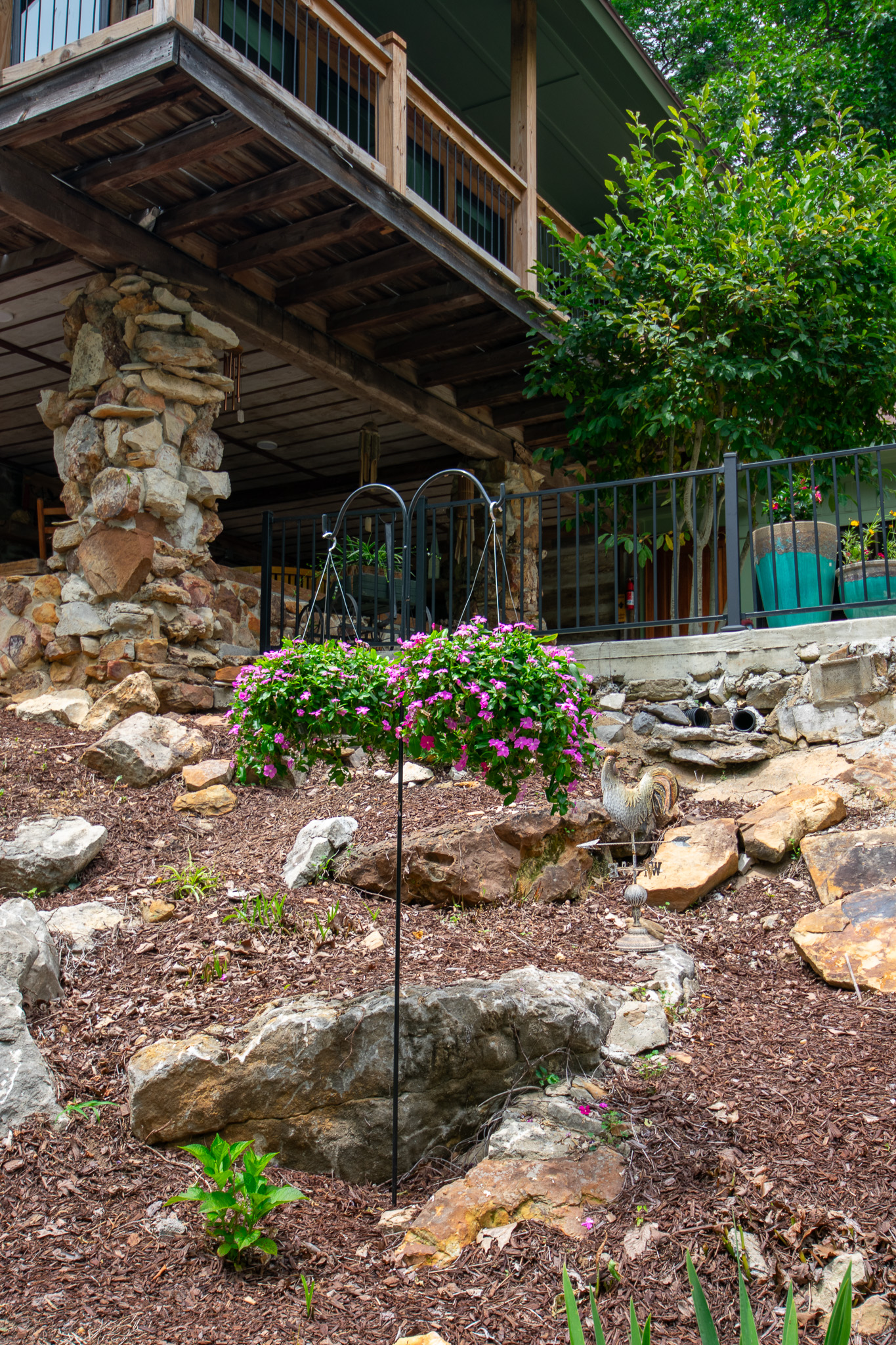 Rocky garden with pink flowers hanging on a black metal stand near a wooden porch and green tree.
