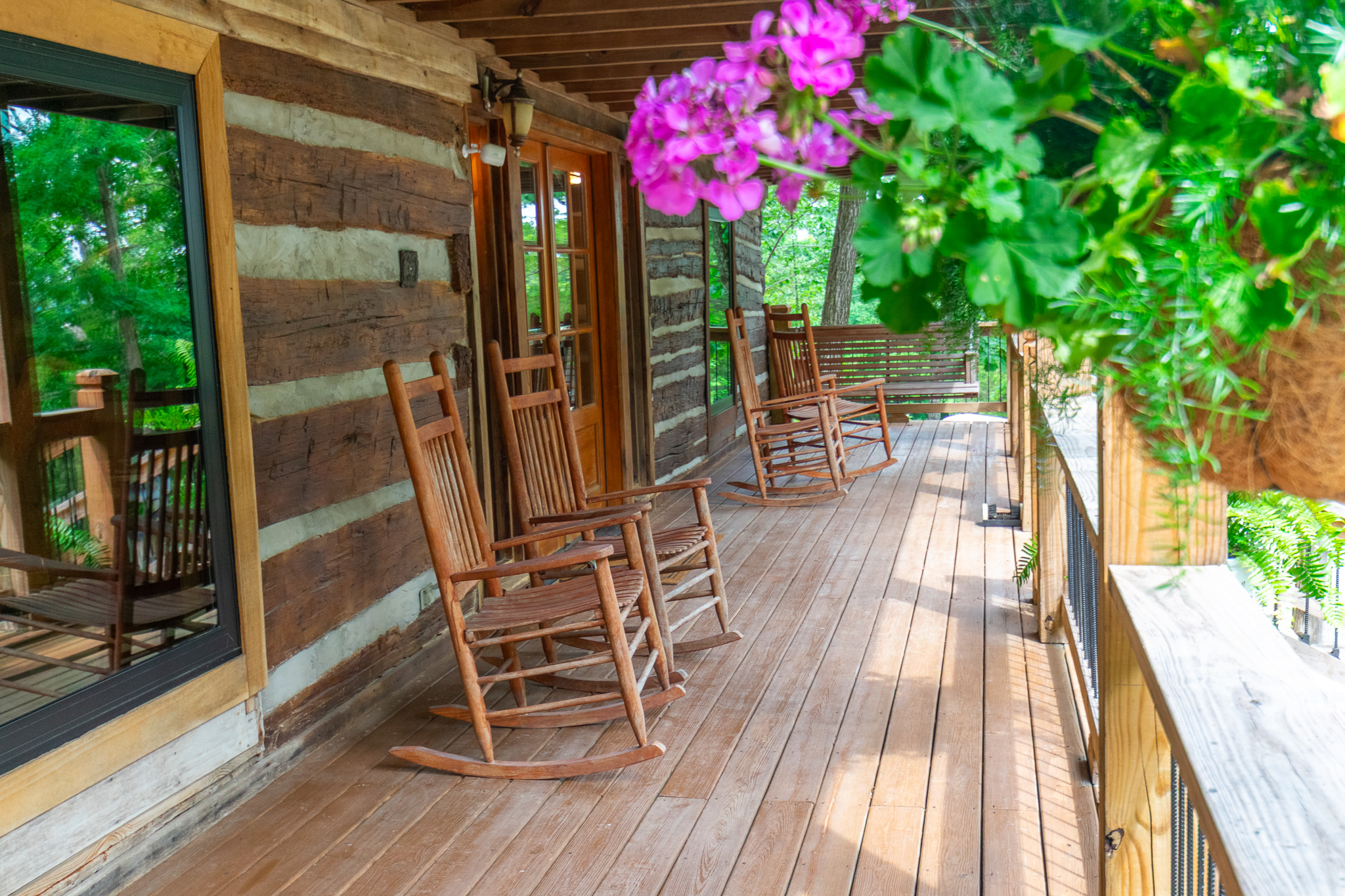 Wooden rocking chairs and a swing on a rustic cabin porch surrounded by green plants and trees.