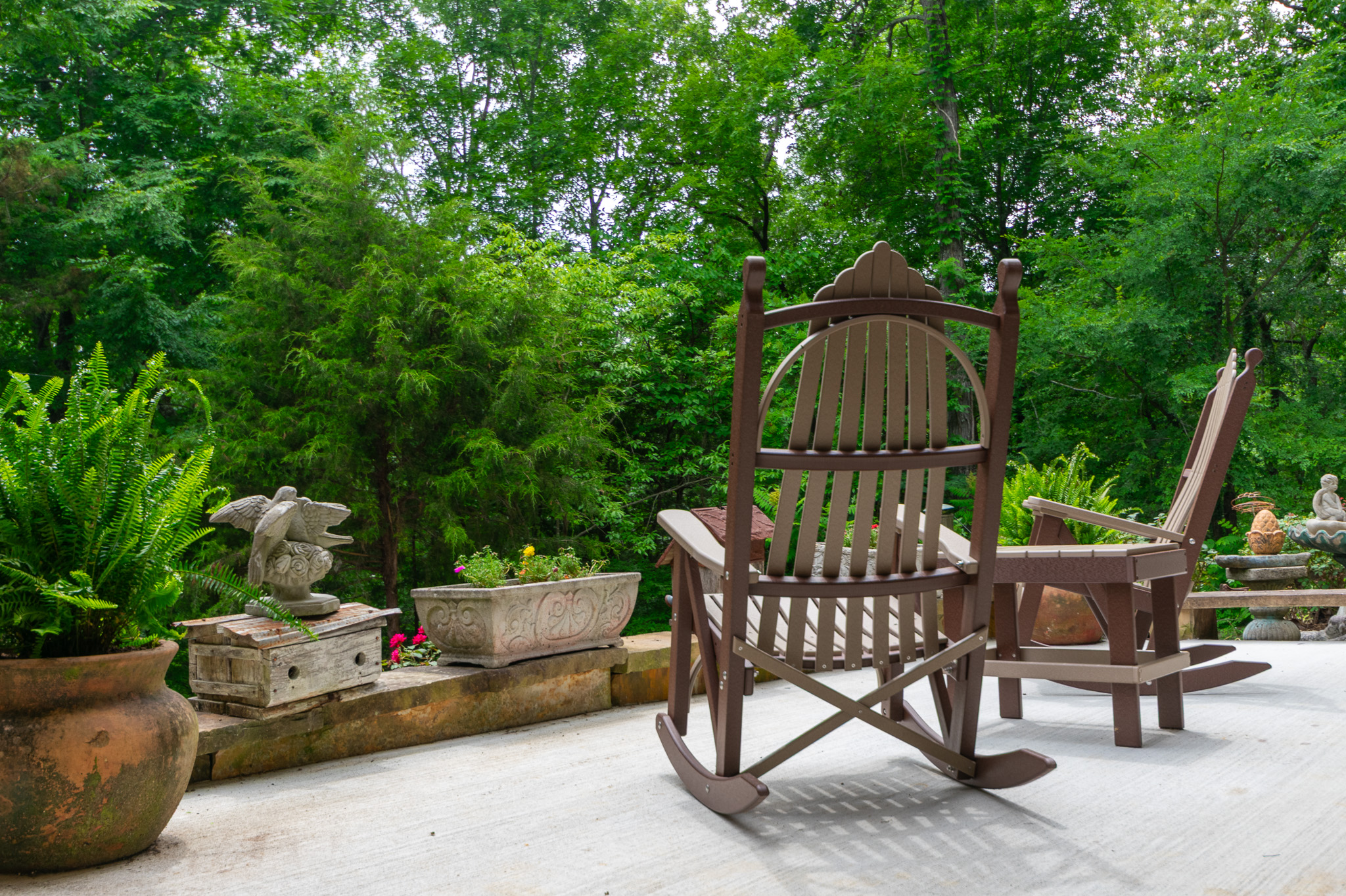 Two brown wooden rocking chairs on a patio with green trees and plants in the background.