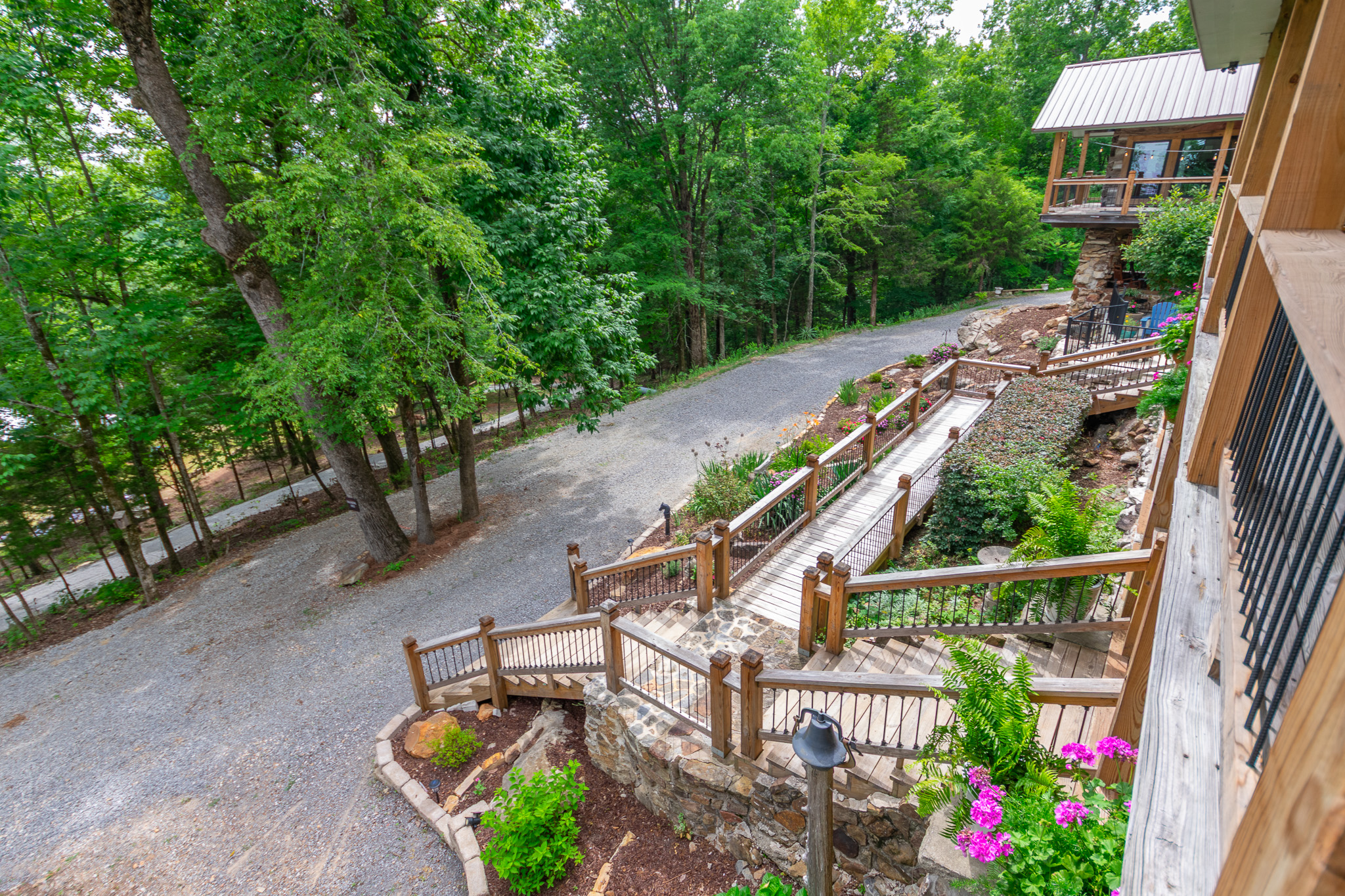 Wooden stairs and bridge with stone wall surrounded by green trees and pink flowers by a gravel path.