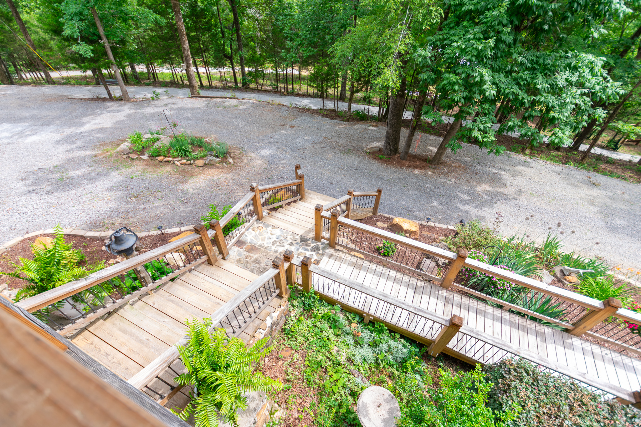 View down wooden steps and ramp leading to gravel driveway surrounded by green plants and trees.