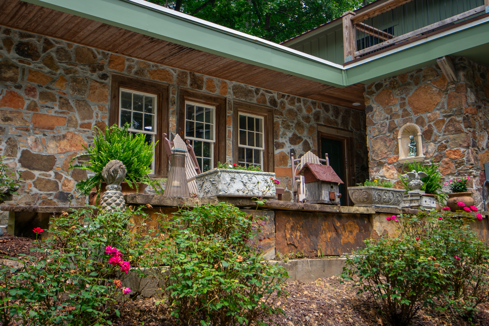 Stone patio with planters, ferns, garden decorations, and pink roses in front of a stone house.