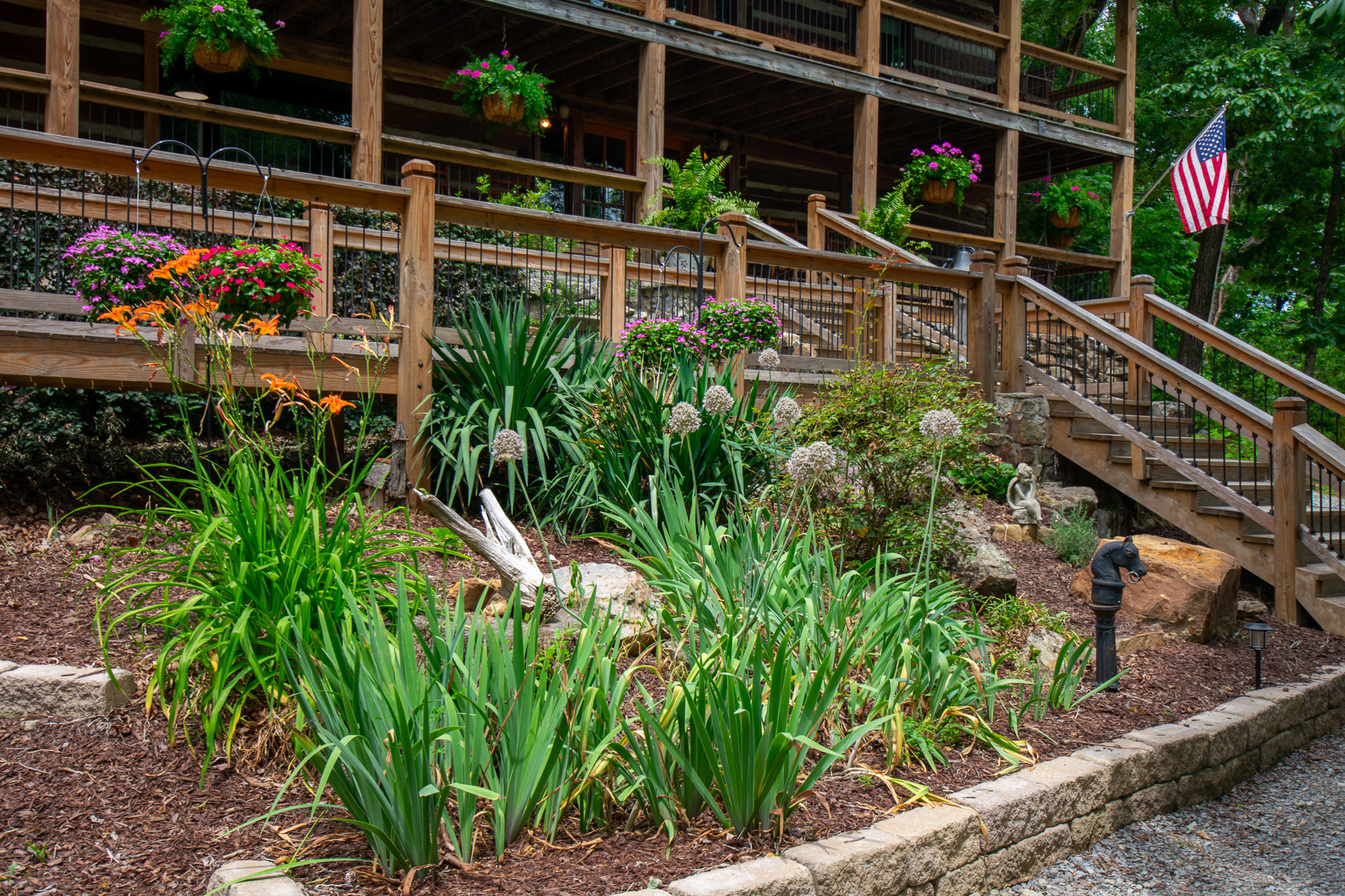 Well-maintained garden with flowers and greenery in front of a wooden porch with hanging plants and stairs.