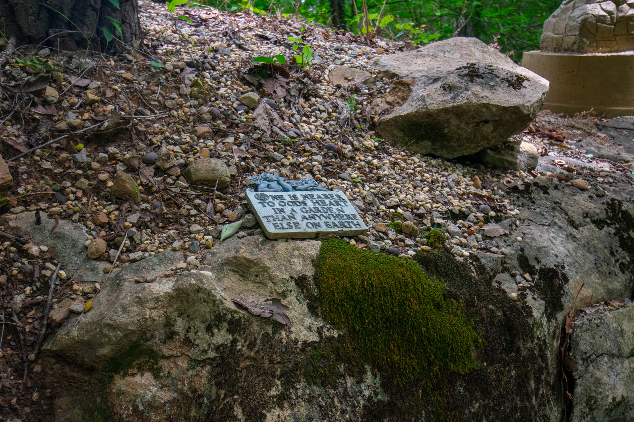 A small garden plaque resting on mossy rocks with gravel and leaves around it in a forest.